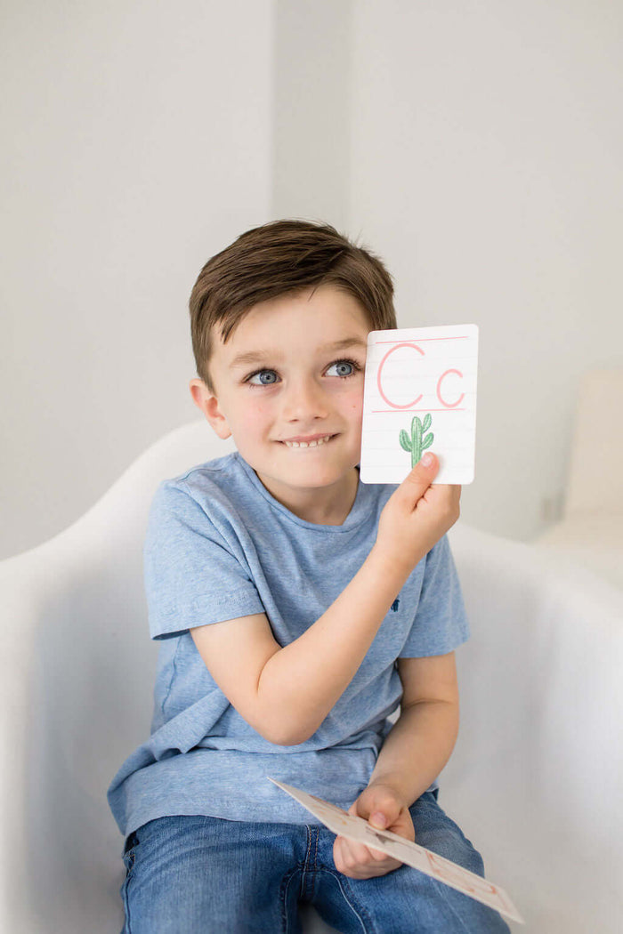 Smiling boy holding a flash card with the letter 'C' and a cactus illustration, learning the alphabet.