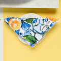 A vibrant, triangular cloth with a colorful floral and lemon pattern sits neatly folded on a round, white plate. The Cocopup London Bandana - Amalfi Lemon features a small label with the text "C O C O P U P London." The background is a contrasting yellow, adding to the cheerful aesthetic of this delightful dog accessory.