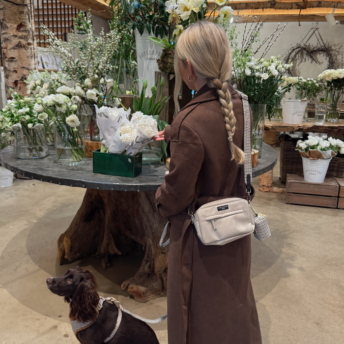 Woman with a braided ponytail shopping for white flowers, dog wearing a check patterned Cocopup London bag strap.