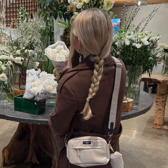 Woman with a braid in a brown coat holds white flowers and carries a Cocopup London bag with Heritage Check strap.
