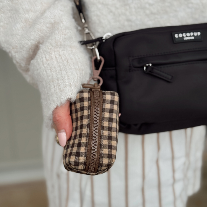 Hand holding a Cotswold Check patterned poop bag holder next to a black crossbody bag, fuzzy sweater visible.