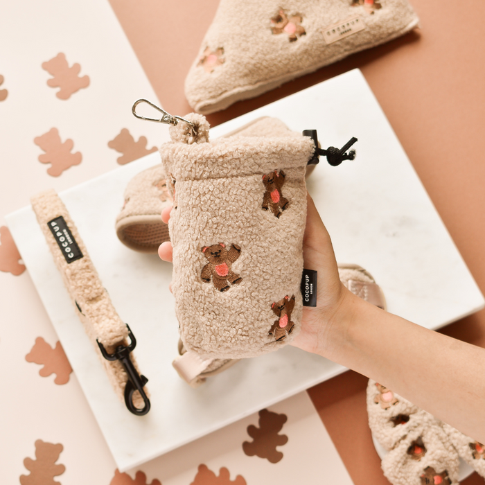 Hand holds a beige Cocopup London drawstring treat pouch with teddy embroidery; matching accessories are nearby.