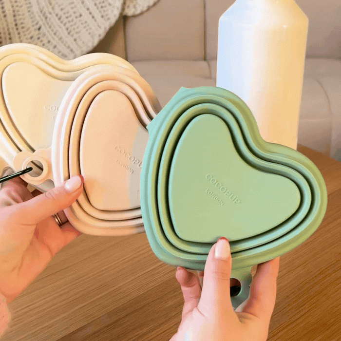 Hands hold a cream Cocopup London heart-shaped collapsible bowl on a wooden table beside a vase.