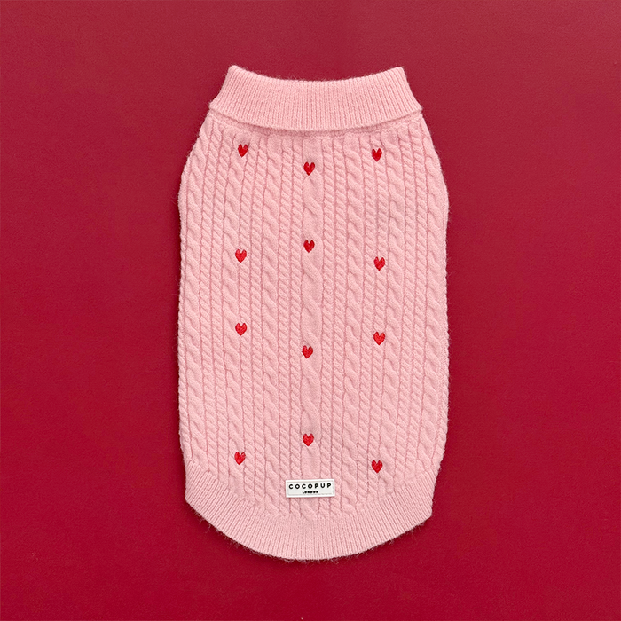 Pink cable knit Cocopup London jumper with heart pattern, displayed on a red background decorated with small red hearts.