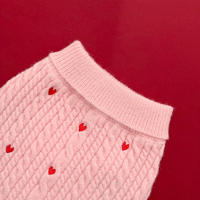 Pink cable knit dog jumper with red heart pattern, ribbed cuffs, set against a red background.