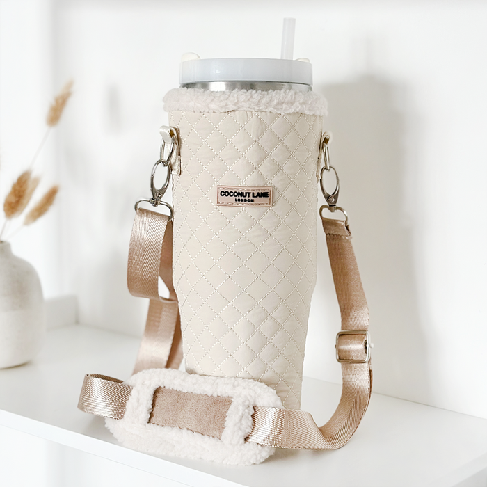 Cream quilted tumbler carry case with beige strap, displayed on a white shelf next to a vase.