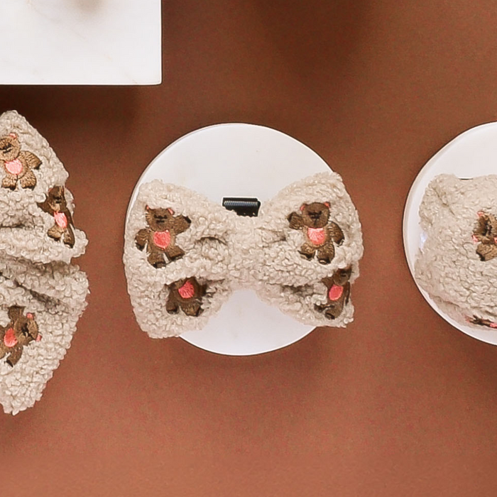 Fuzzy beige teddy bow tie with pink hearts, embroidered, displayed on a round white card.