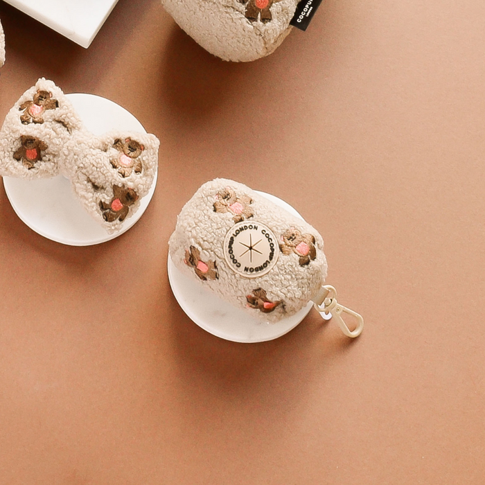 Cocopup London Theo Embroidered Teddy Poop Bag Holder in soft beige displayed on white dishes on a light brown surface.
