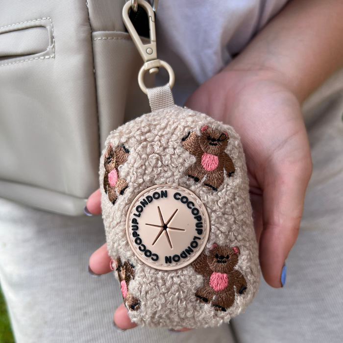 Brown teddy bear-shaped embroidered poop bag holder attached to a bag, held by a hand.