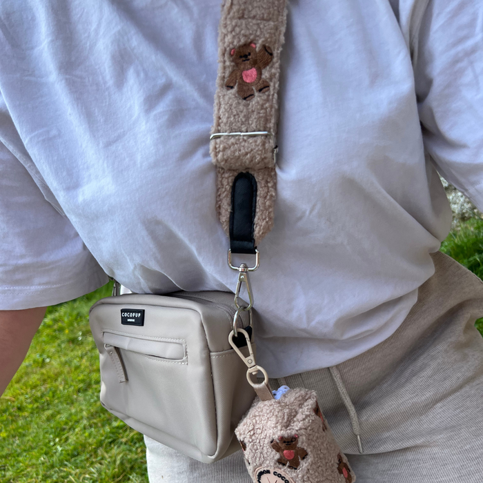Person in a white shirt and beige pants holding a beige Cocopup London Teddy poop bag holder with embroidered details.